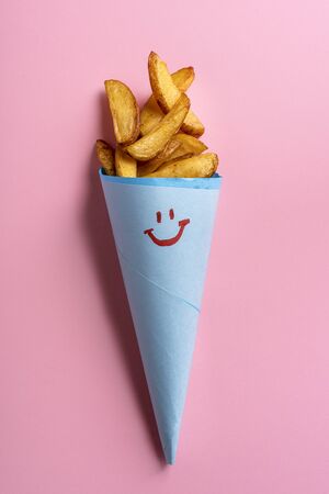 Baked potatoes wedges in a blue paper cone with a smiley face on a pink background. Tasty roast potatoes in a cone. Homemade rustic potatoes.の写真素材