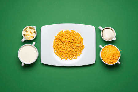 Uncooked pasta and the ingredients for making mac and cheese. White plate with raw macaroni on green...