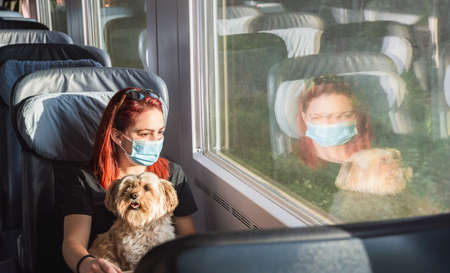 Young redheaded woman with face mask traveling by train, at first-class. Girl and dog travel by german intercity express train. Pandemic safe travel.の写真素材