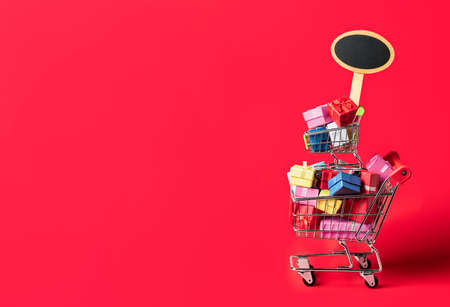 Shopping cart full of colorful gifts and an empty placard, isolated on a red background. Black...