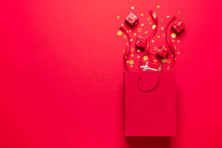 Flat lay with golden confetti, red shopping bag and gift boxes on a red-colored background. Gift boxes coming out of a red paper bagの写真素材