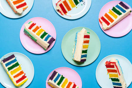Slices of rainbow cake on colorful plates, isolated on blue background. Flat lay with buttercream...