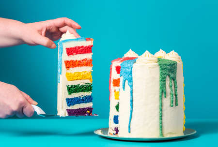 Woman's hands taking a slice of rainbow cake. Delicious homemade birthday cake against a blue background.の写真素材