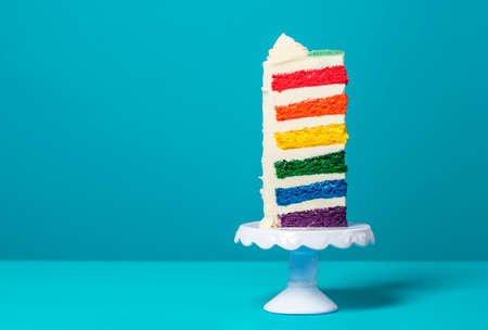 Single slice of birthday cake on a white cakestand minimalist on a blue background. Homemade cake with multicolored layers and buttercreamの写真素材