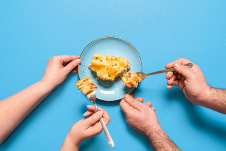 People sharing and eating a slice of lasagna, isolated on blue background. Flat lay with vegetable...