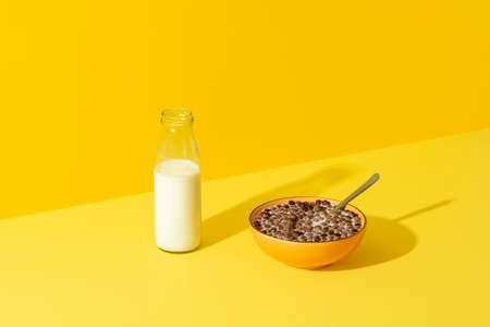 Bowl with cereals and a glass bottle of milk on a yellow table in bright light. Breakfast table with...