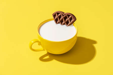 Single yellow cup of milk and a waffle as breakfast isolated on a colored table. Milk and heart-shaped waffle minimalist on a yellow background.の写真素材