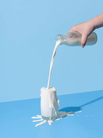 Woman hand pouring milk from a bottle into the glass, minimalist on a blue background. Over-spilling milk on the blue-colored table.の写真素材