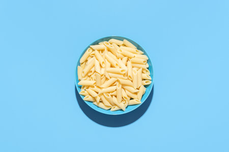 Above view with a penne pasta bowl minimalist on a blue table. Plain cooked pasta, without any sauce, in a blue bowl.の写真素材