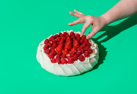 Woman's hand grabbing a raspberry from a pavlova cake, minimalist on a green background. Homemade pavlova cake with whipped cream filling, strawberries, and raspberries on top of it.の写真素材