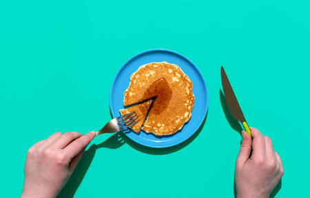 Above view with woman's hands slicing and eating a pancake. Pancake with maple syrup on a blue plate isolated on a green backgroundの写真素材