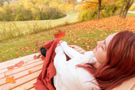 Close-up with a young woman sitting on a bench covered with an orange blanket. Woman enjoying a beautiful day of autumn in the parkの写真素材