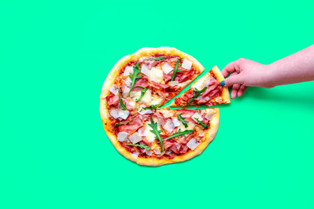 Top view with a homemade ham pizza isolated on a green background. Woman's hand taking a slice of pizzaの写真素材