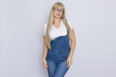 Sweet beautiful blond woman in dark jeans overalls and glasses over grey backgroundの写真素材