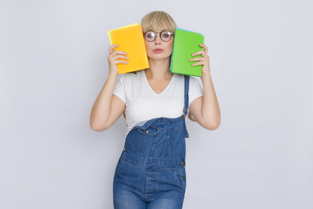 Studio portrait of a beautiful young blonde woman in blue denim overalls and glasses holding books...