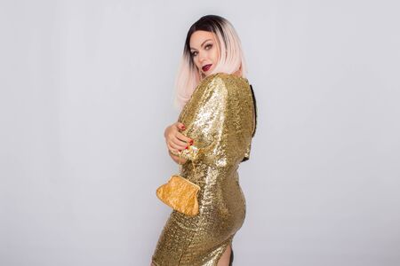 Charming young blonde haired woman wearing elegant gold evening dress and holding gold color clutch...