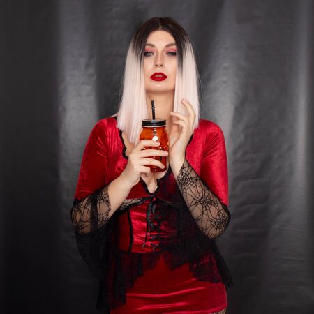 Young beautiful blonde woman in a red vampire dress holds an orange glass in the shape of a skull in...