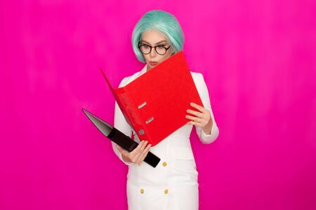 Beautiful young woman with short blue hair wearing glasses in a white business jacket dress holds two thick folders in her handsの写真素材