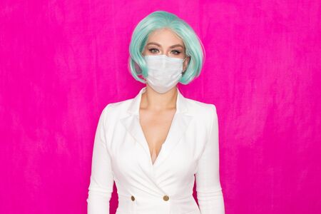 Beautiful young woman with short blue hair in a white business dress jacket with a medical mask posing on a pink background in the studioの写真素材