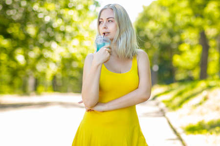 attractive stylish blond smiling woman in yellow short dress, summer fashion outfit, drinking cocktail in parkの写真素材