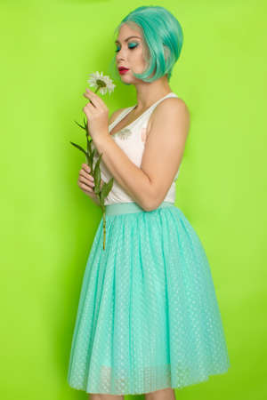 Studio portrait of beautiful mint hair feminine in white top and light blue skirt holding big chamomile in her hands, over green backgroundの写真素材