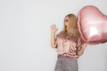 Blonde business woman in pink blouse holding big pink heart balloon in her hands over white...
