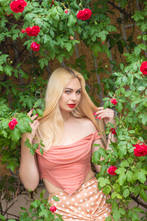 Beautiful woman with long blonde hair and red lips wearing pink clothes poses near blooming roses in garden. Wear light pink top and skirtの写真素材