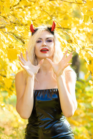 Beautiful blonde woman in a devil costume in nature. Black latex tight dress and horns. Soft focus. Halloween conceptの写真素材