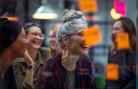 Women in their thirties and an older woman laughing, brainstorming around a glass board with notesの素材