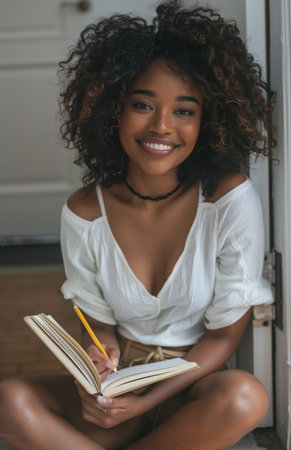 Beautiful black woman sitting on the floor of her hallway holding an open notebook and pencilの素材