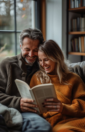 A joyful couple relaxes on the sofa at home, reading a book and laughing togetherの素材