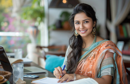 An Indian woman in a saree writing on a notepad at a table with a laptop and waterの素材