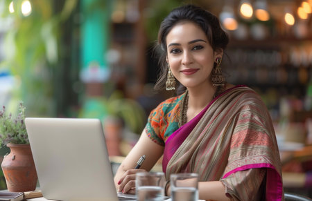 An Indian woman in a saree writing on a notepad at a table with a laptop and waterの素材