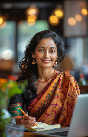 An Indian woman in a saree writing on a notepad at a table with a laptop and waterの素材