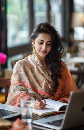 An Indian woman in a saree writing on a notepad at a table with a laptop and waterの素材