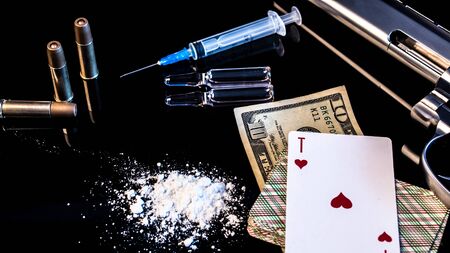 A pistol with cartridges, syringes, ampoules, white powder and playing cards lie on a black glass...