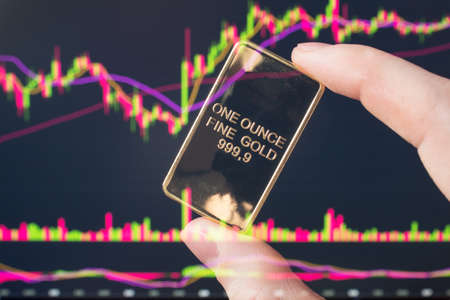 The hand holds a gold bar on the background of a laptop on which there is a trading price chart. One ounce of gold in hand. Trading gold on the stock exchange and Forex.のeditorial素材
