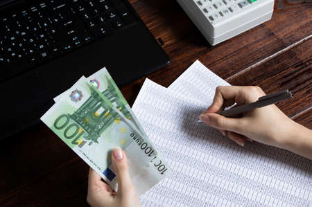 Obligation to pay wages and debts in the company.A cashier holds money Euro over an office work space with documents, a cash register,a phone,and a computer.Work in the office with finances in Europeのeditorial素材