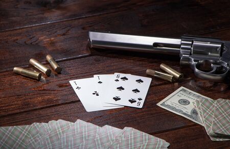 Gambling in cards, poker or blackjack for money. On a wooden table are a pistol with cartridges, cash dollars, and a deck of playing cards with an ACE of spades and a six. Dangerous gambling debtsの写真素材