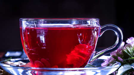 A mug of red tea in white hibiscus flowers and green leaves of medicinal tea on a wooden stand.Zen tea ceremony. Photo of red herbal Indian healing tea. Elegant mugs with a relaxing and tonic drinkの写真素材