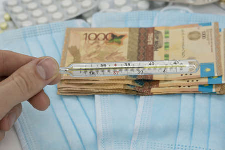 A hand holds a thermometer with a high temperature over a table with pills, a medical mask and tenge...