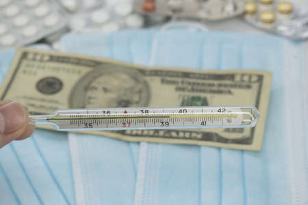 A hand holds a high temperature thermometer over a table with pills, a medical mask and dollar money. The growth of prices for medicines in America in dollars. The United States is under quarantineの写真素材