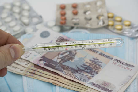 A hand holds a thermometer with a high temperature over a table with pills, a medical mask and money...