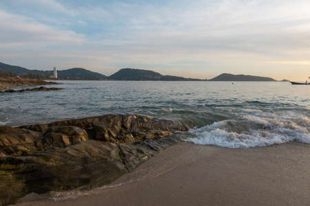 A view of a sandy beach with a large stone on the shore about which waves are beatingの写真素材