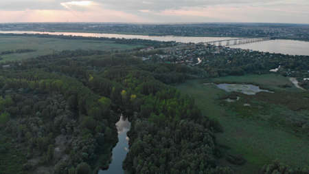 Aerial view of a green forest with a small river. Automobile bridge over the Dnieper river in the background. There is a city on one bank of the river and summer cottages on the other bank.の写真素材
