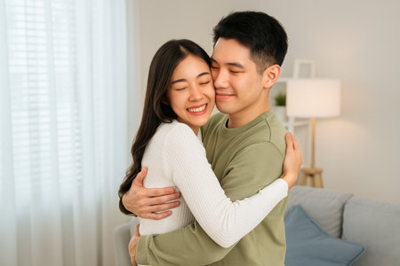 A young couple smiles as they share a warm hug in a cozy living room. Sunlight streams through the curtains, creating a joyful and intimate atmosphere.の素材