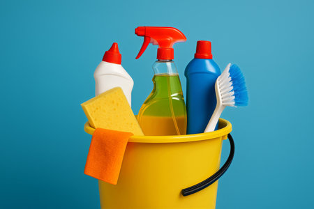 A bright yellow bucket contains various cleaning supplies including sponges, sprays, and cleaning solutions.の素材