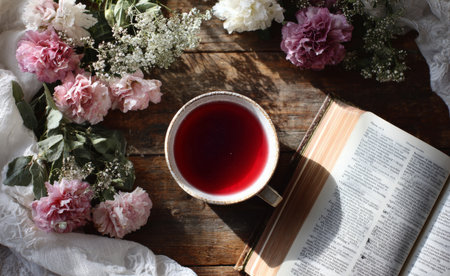 A warm cup of red tea sits on a rustic wooden table surrounded by colorful flowers and an open book. Soft natural light creates a cozy atmosphere, perfect for reading.の素材