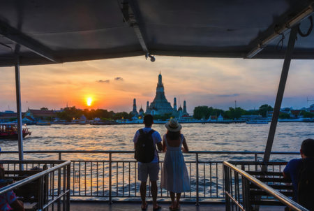 Over-the-shoulder view of foreign travelers on a Thai ferry looking toward Wat Arun at sunsetの素材