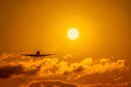 Silhouette of airplane flying into sunset sky with clouds and glowing sun. Airplane Silhouette at Sunset Sky with Glowing Sun and Clouds.の写真素材